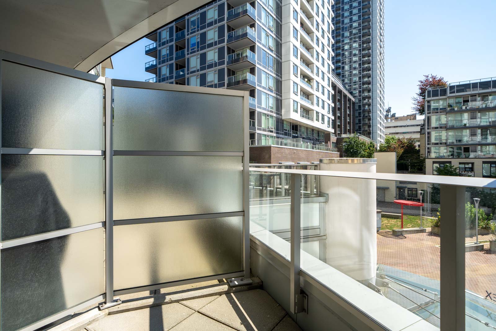 View from a modern apartment balcony with frosted privacy panels, glass railing, and high-rise buildings in the background on a sunny day.