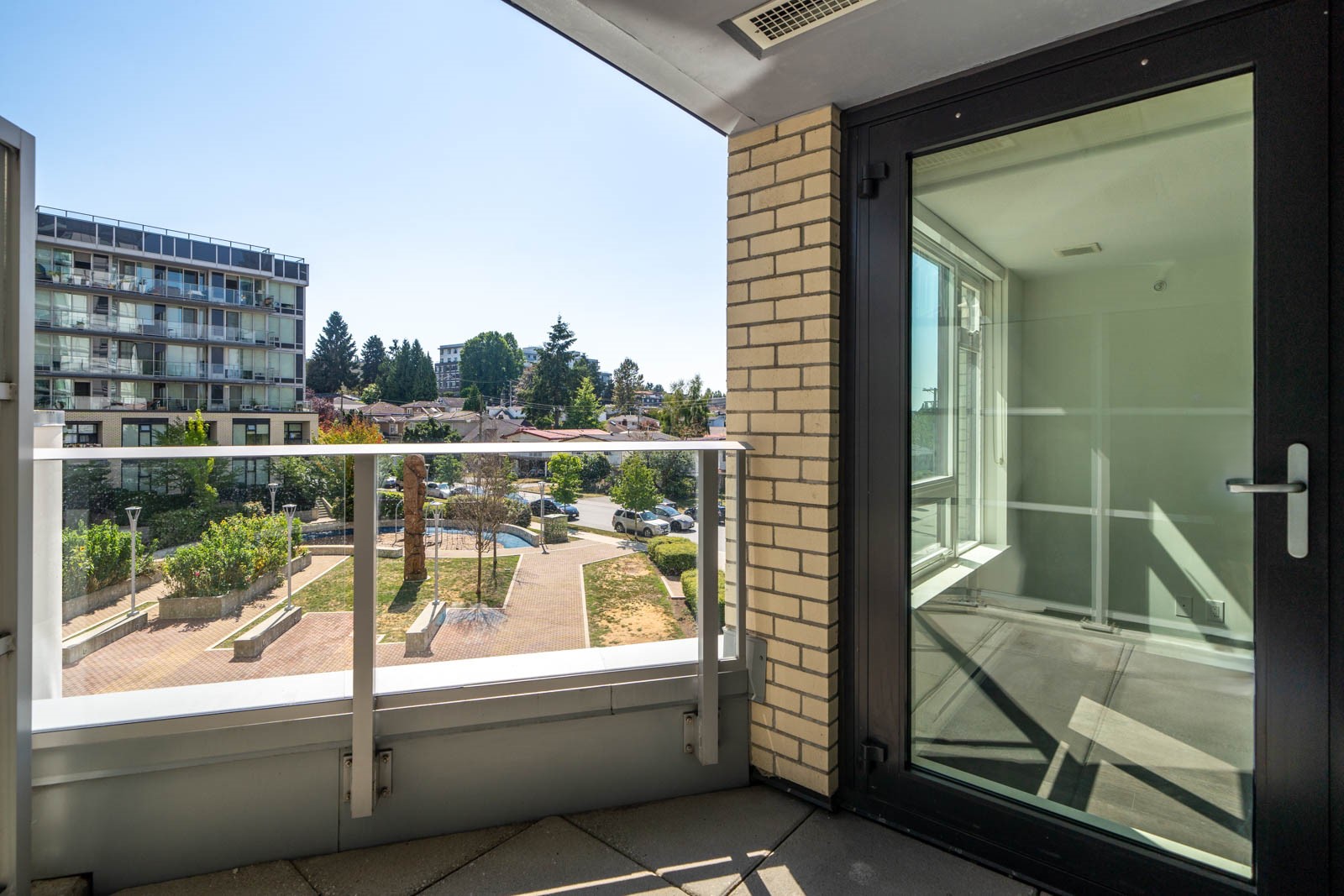 A modern apartment balcony with glass railing overlooks a small park and residential buildings on a sunny day. A glass door leads inside to an empty room.