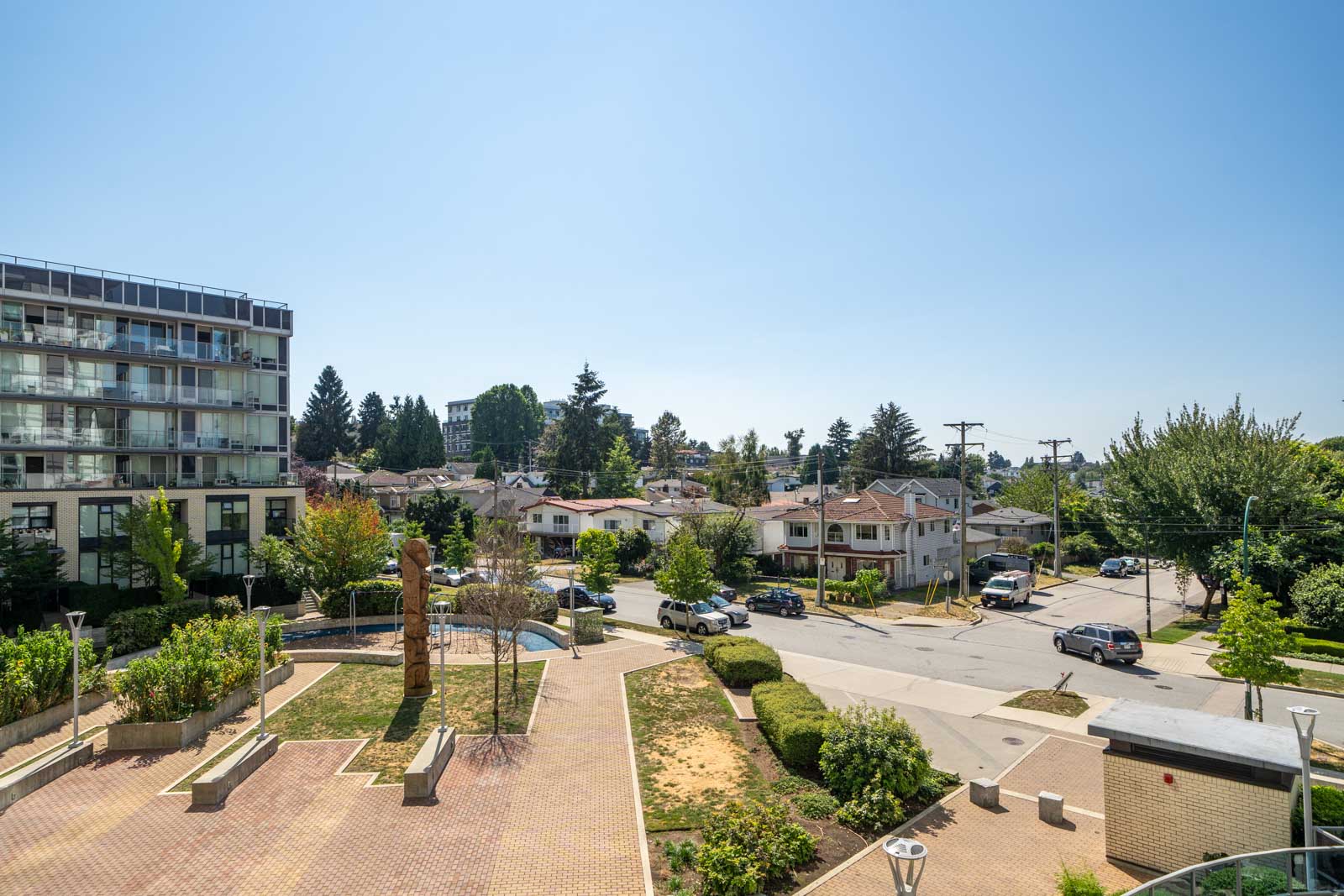 View of a residential neighborhood with houses, a mid-rise apartment building, parked cars, trees, and a paved courtyard under a clear blue sky.