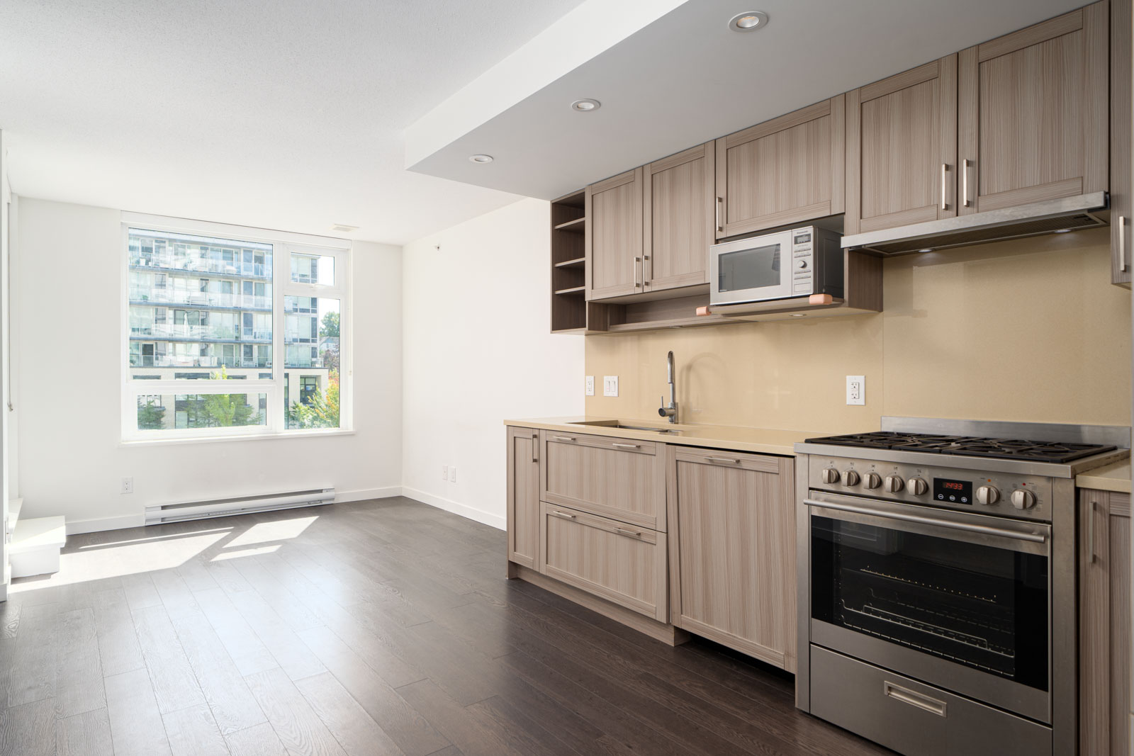Modern apartment kitchen with light wood cabinets, stainless steel oven, built-in microwave, and a large window letting in natural light.