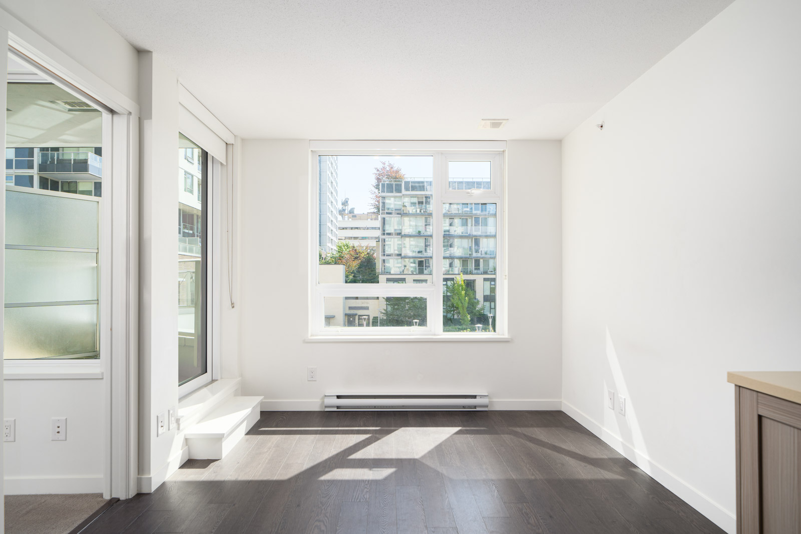 Bright, empty apartment room with large window, dark wood floors, white walls, and a view of modern buildings outside.
