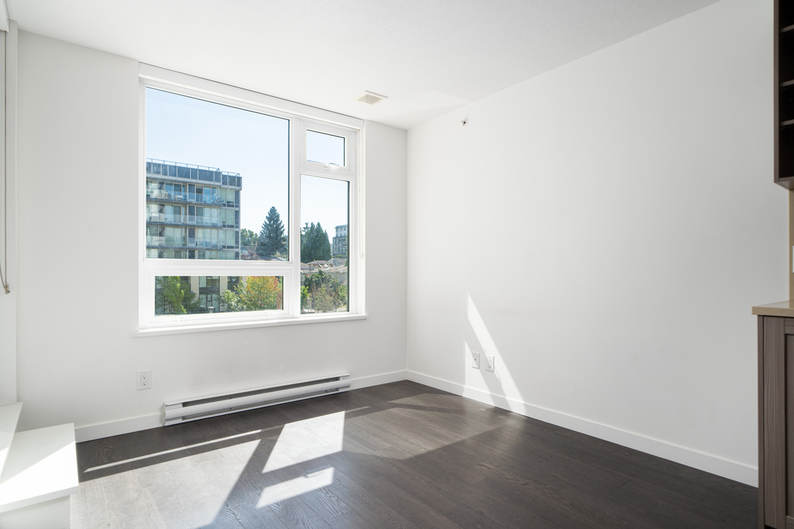 Empty room with white walls, large window letting in natural light, dark hardwood floor, and a view of trees and an apartment building outside.