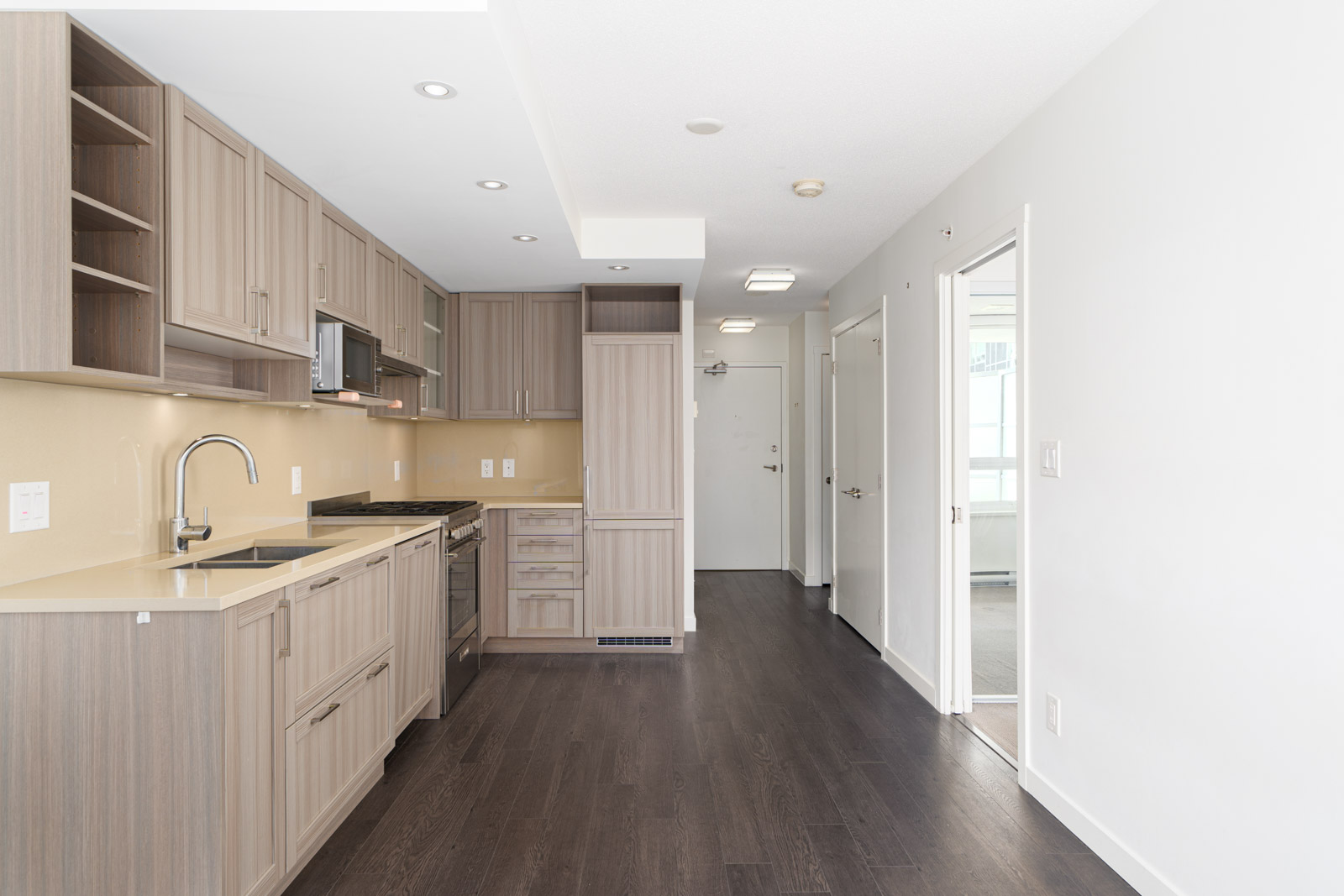 Modern apartment kitchen with light wood cabinets, stainless steel appliances, dark flooring, and open entryway leading to other rooms.