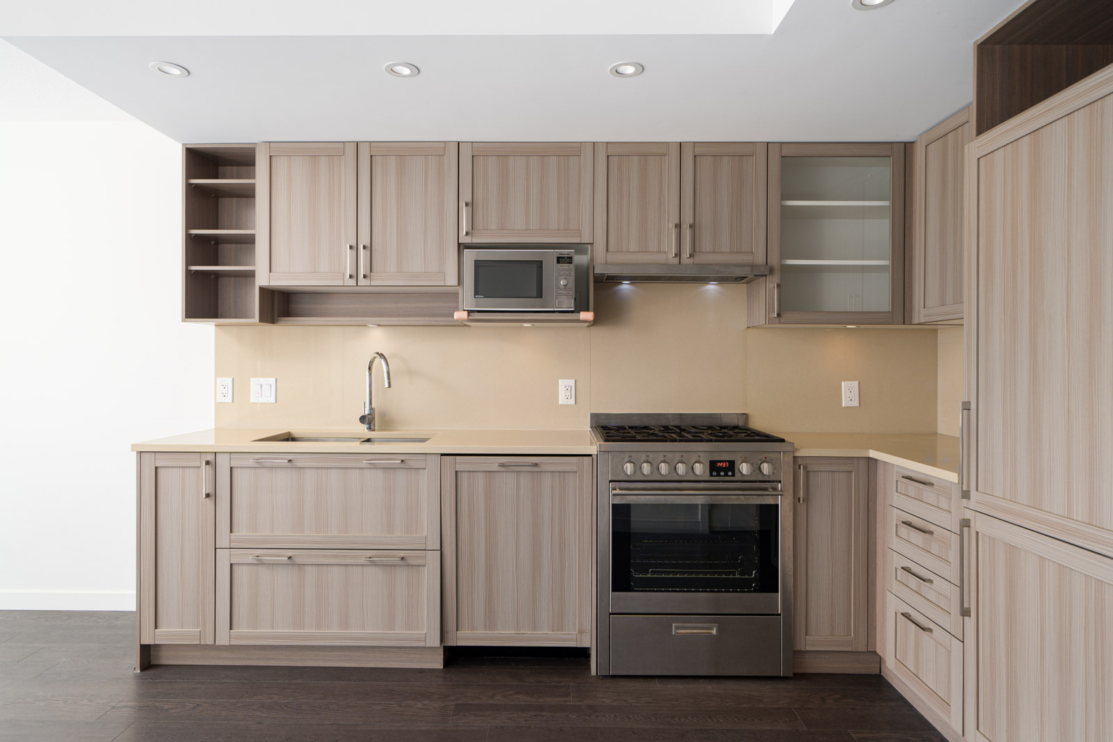 Modern kitchen with light wood cabinets, built-in microwave, stainless steel oven, integrated refrigerator, sink, and beige countertops.