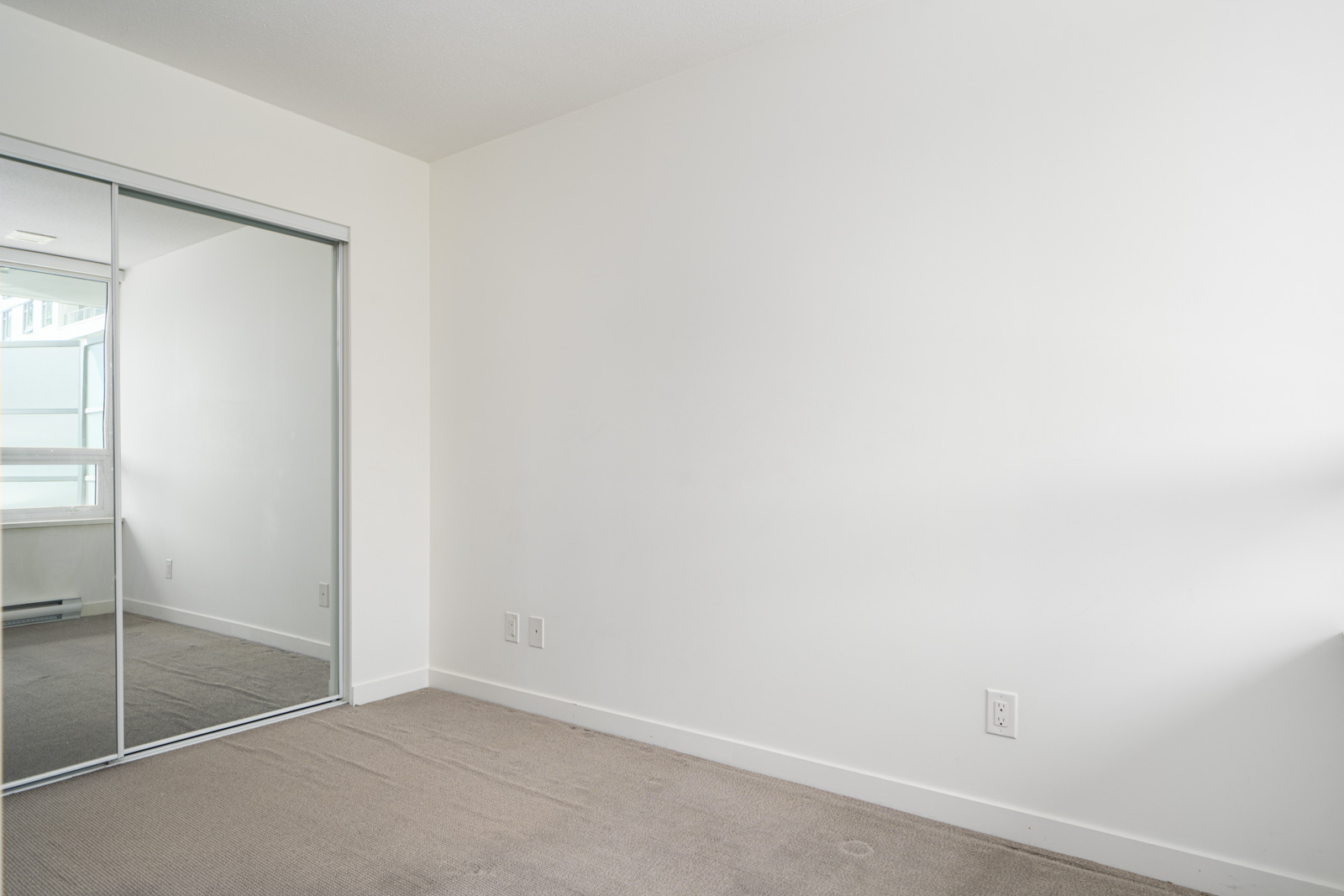Empty room with white walls, beige carpet, a mirrored sliding closet door, electrical outlets, and natural light coming from a window on the left.
