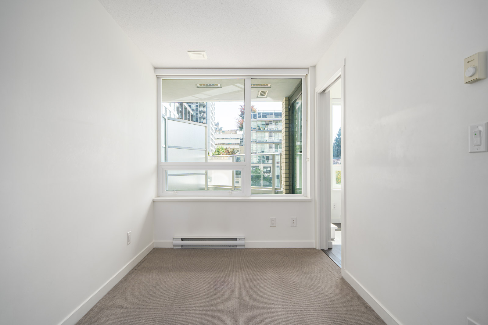 Small, empty room with white walls, beige carpet, large window, and glass door leading to a balcony with views of neighboring buildings.