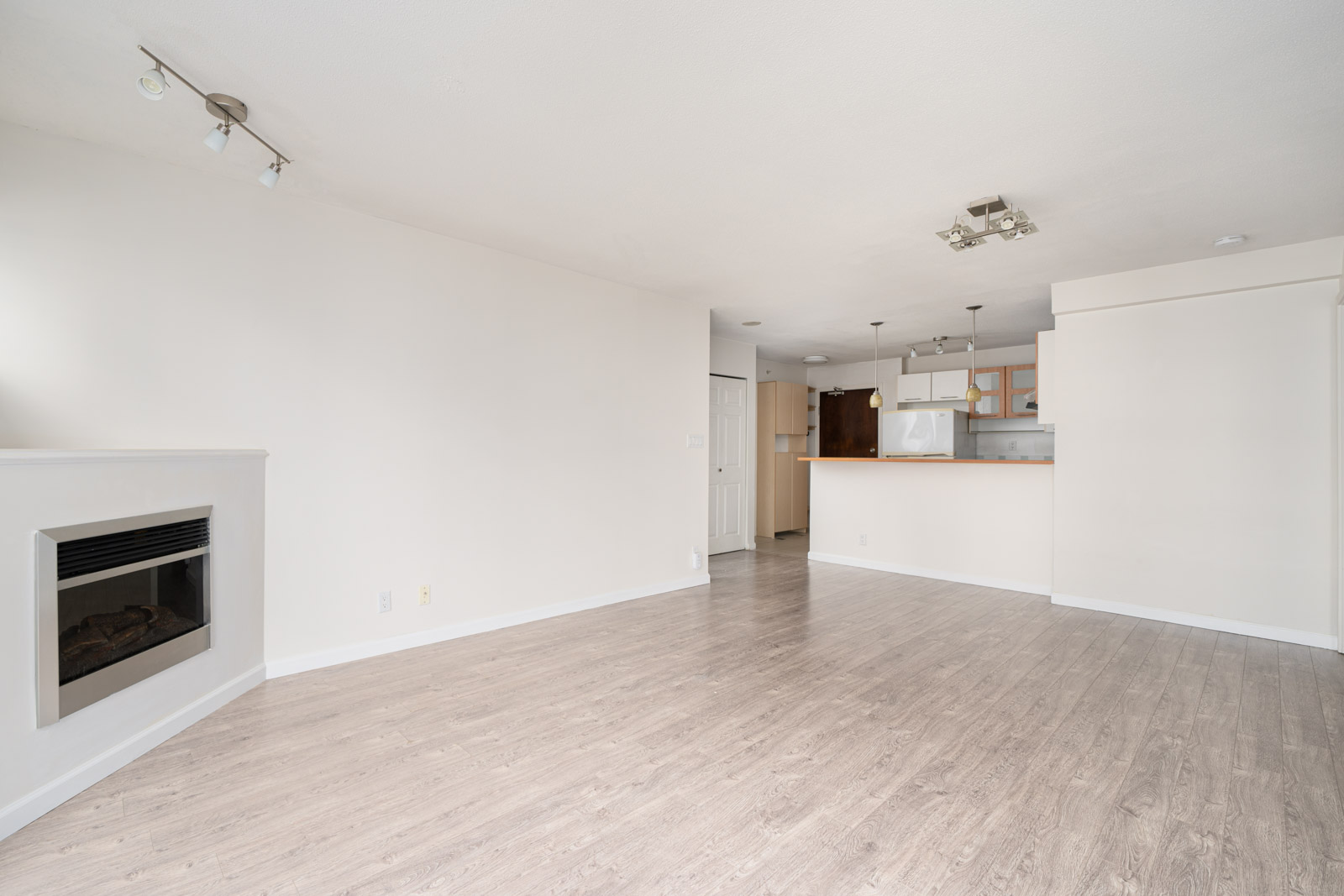 Unfurnished apartment living area with light wood flooring, a built-in fireplace, and an open kitchen with white cabinets and hanging lights.