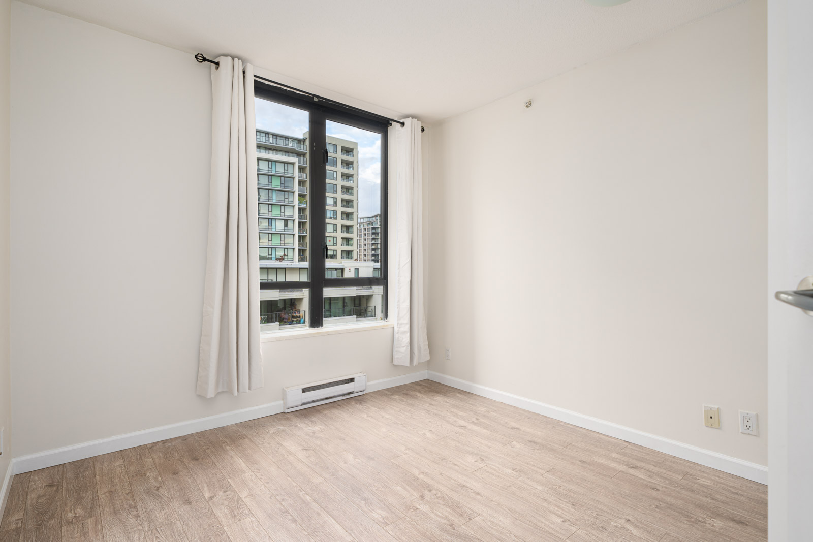Empty room with light-colored walls and wood flooring, featuring a large window with white curtains and a view of modern apartment buildings outside.