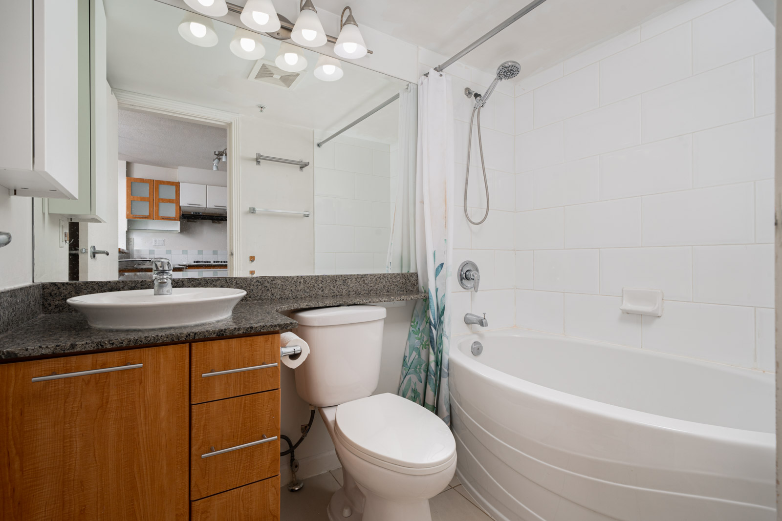 Modern bathroom with a bathtub, showerhead, toilet, vessel sink, wooden cabinet, granite countertop, and large mirror under bright vanity lights.