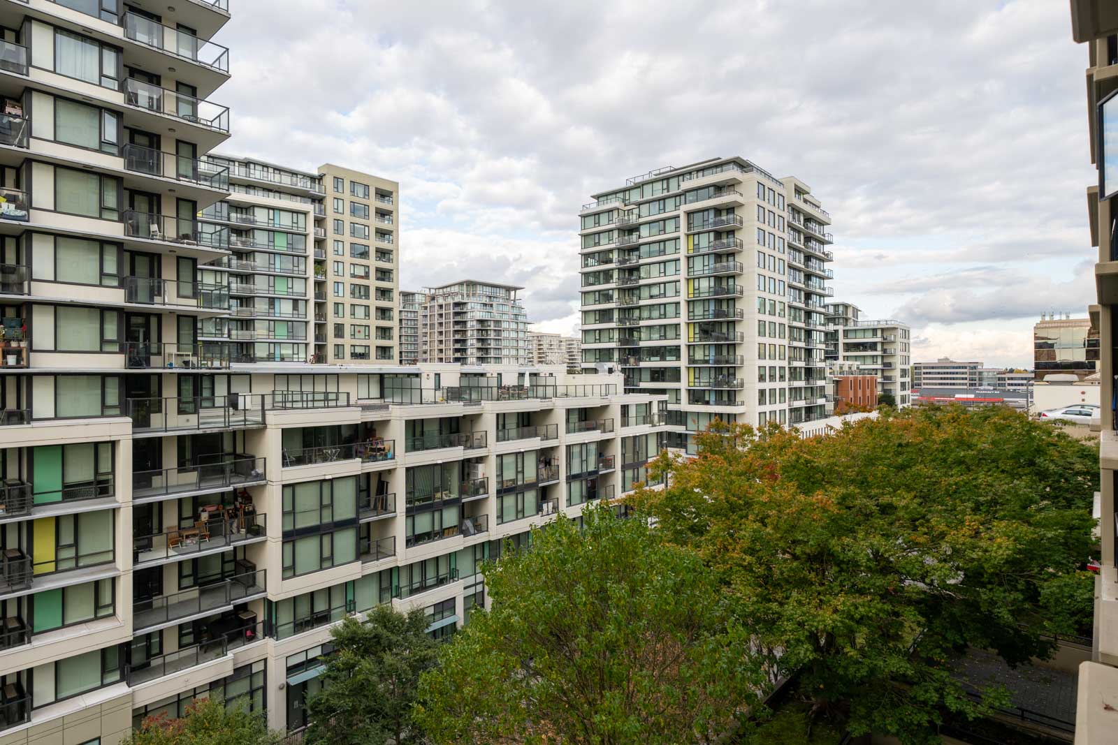 Modern mid-rise apartment buildings with balconies surround a courtyard area filled with green trees under a cloudy sky.