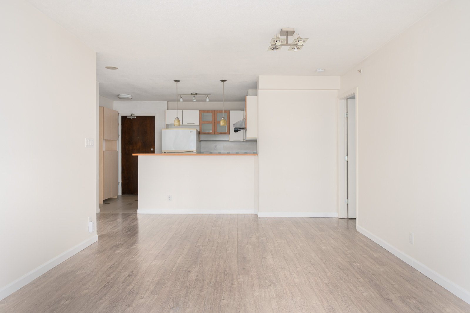 Unfurnished apartment interior with light-colored walls, wood flooring, and an open kitchen featuring white cabinets and appliances.