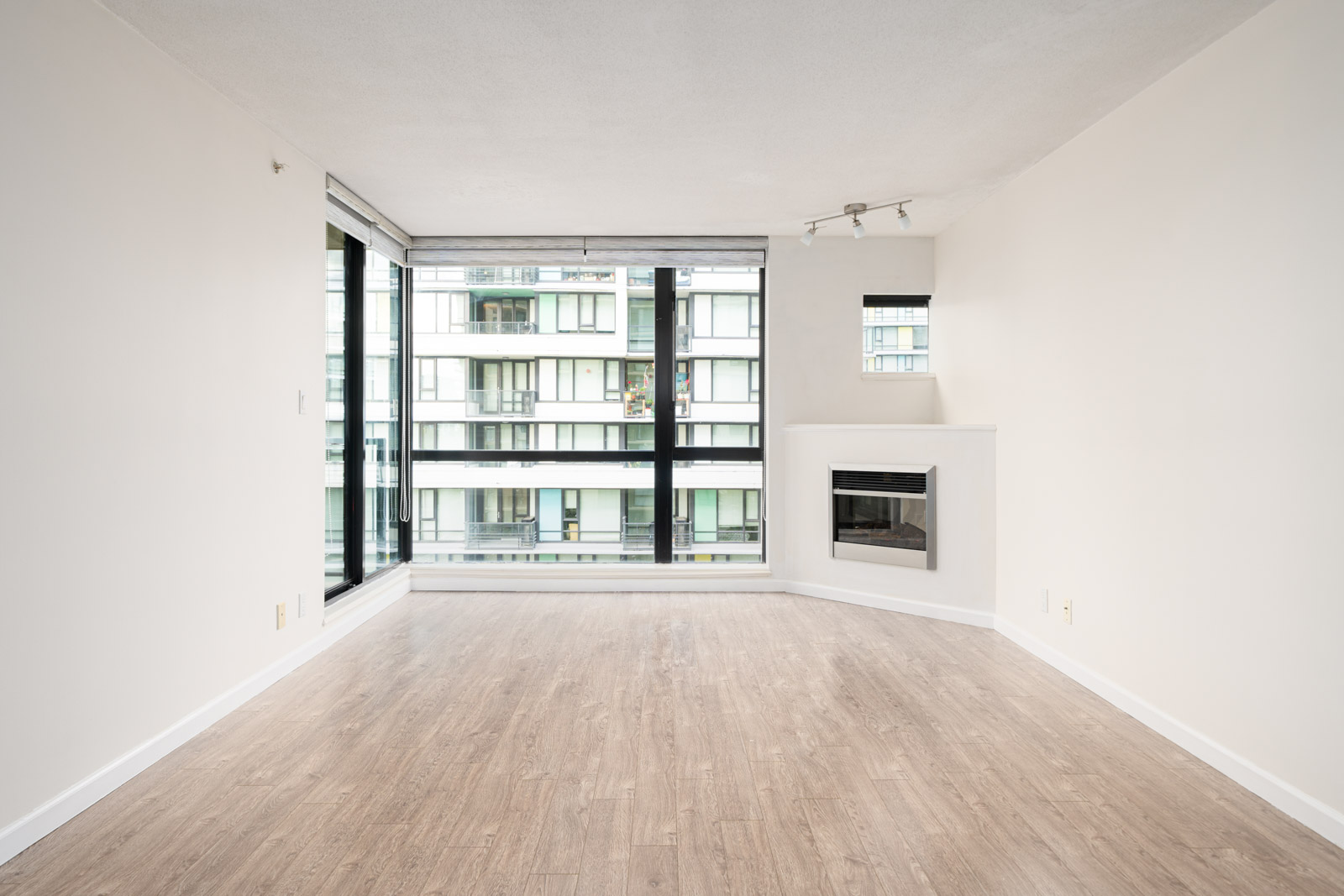 Empty living room with light wood flooring, large floor-to-ceiling windows, a white wall, and a built-in fireplace; exterior apartment building visible outside.