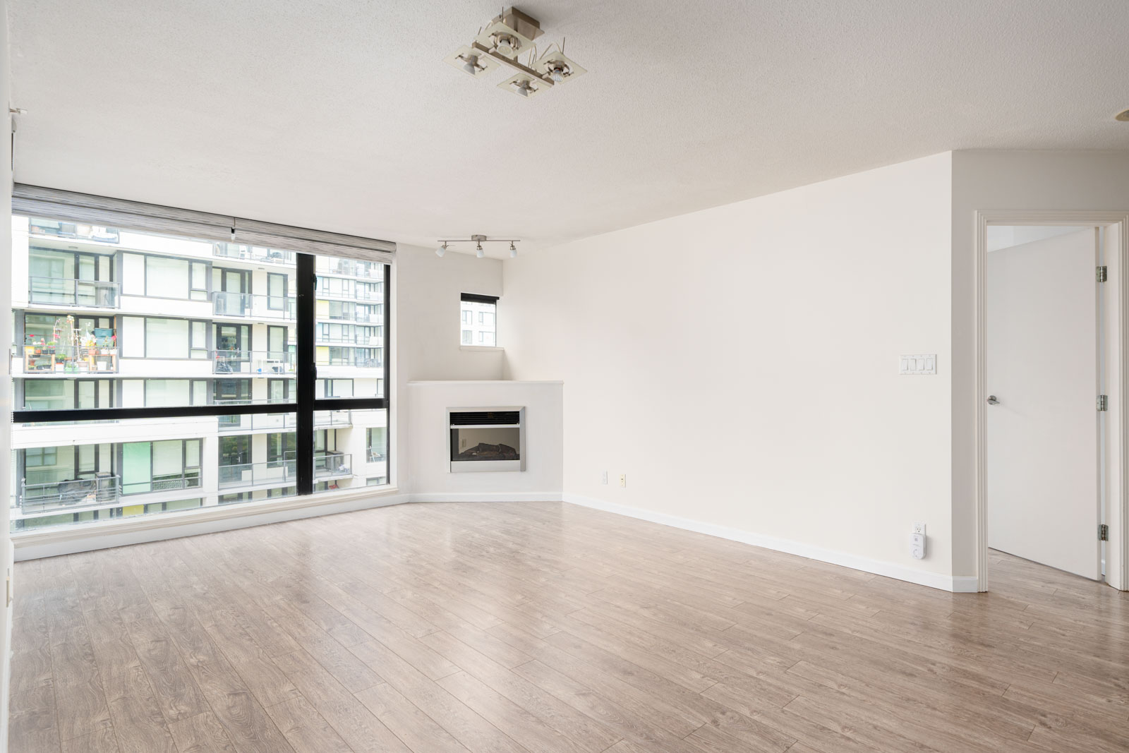 Unfurnished living room with large windows, light-colored walls, wood flooring, a small fireplace, and an open doorway leading to another room.