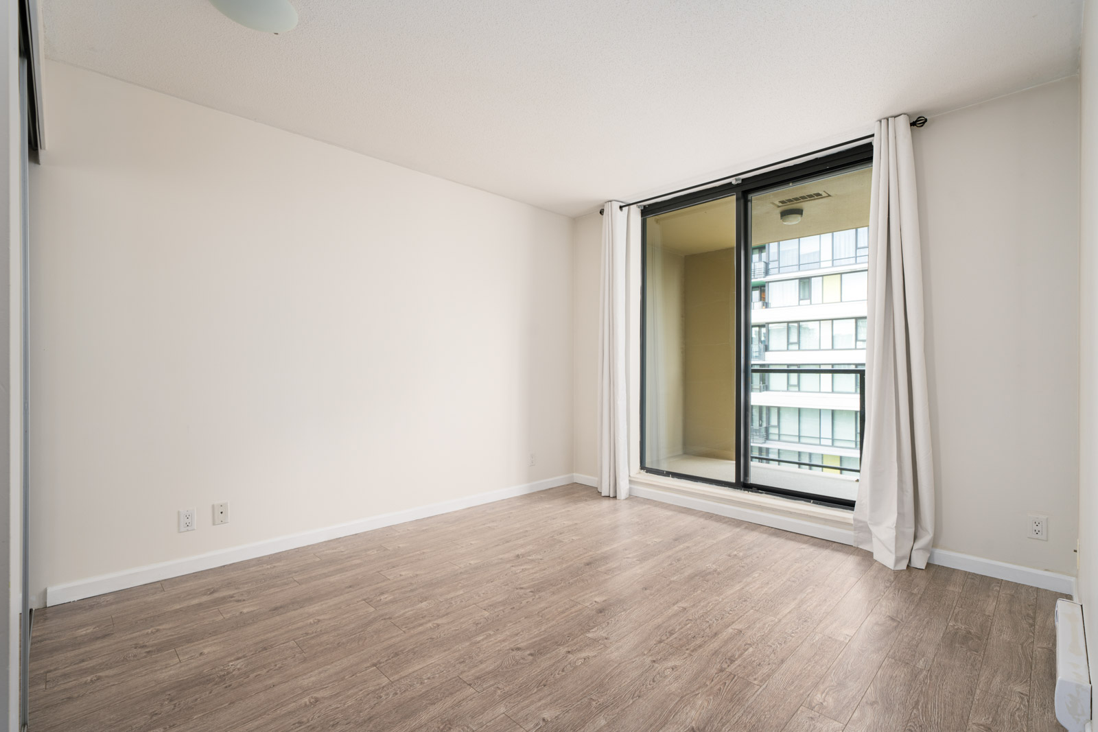 Empty room with light wood flooring, white walls, a ceiling light, and large glass doors leading to a balcony with white curtains. A modern building is visible outside.
