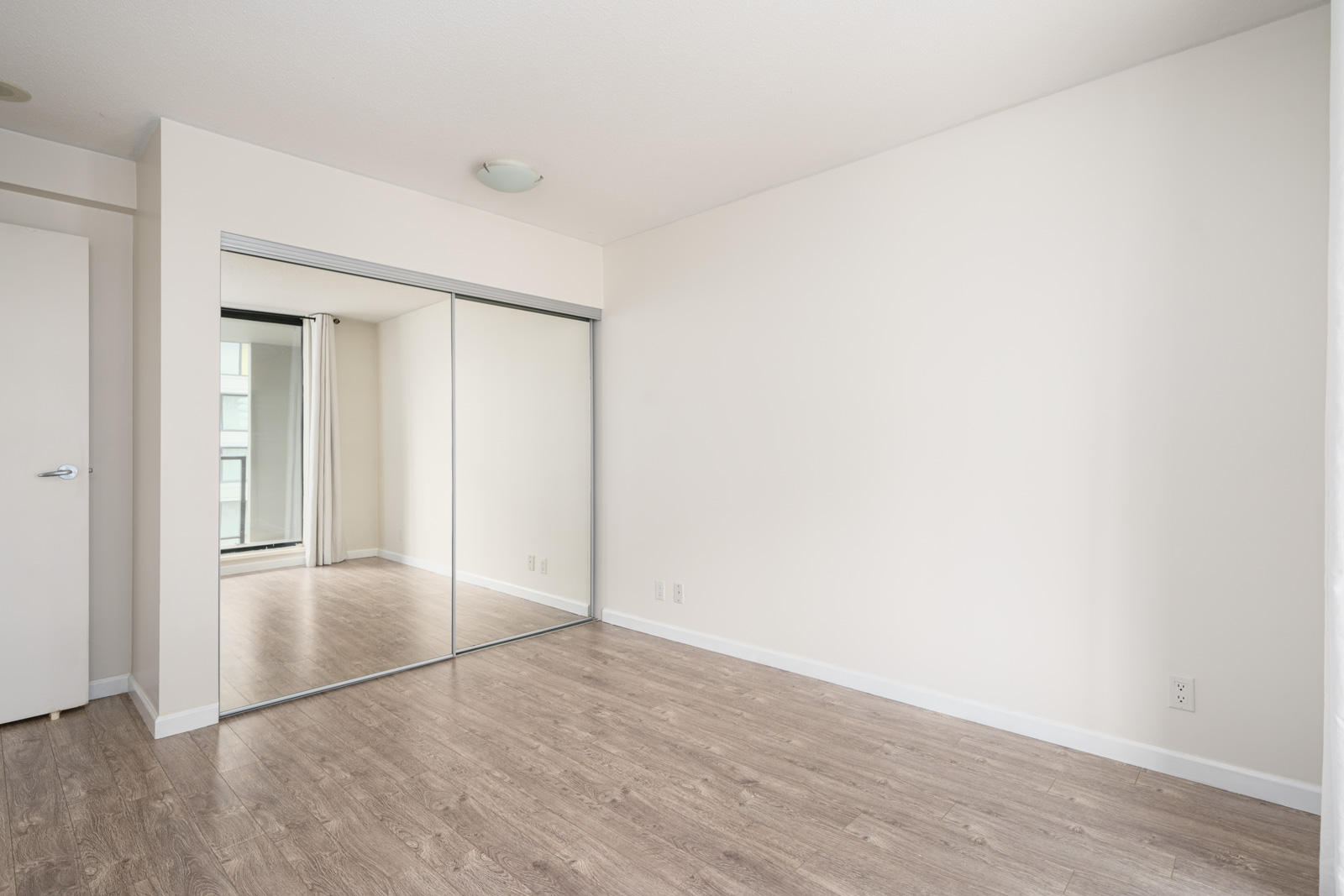 Empty room with light-colored walls, a hardwood floor, large sliding mirrored closet doors, and a window reflected in the mirror.