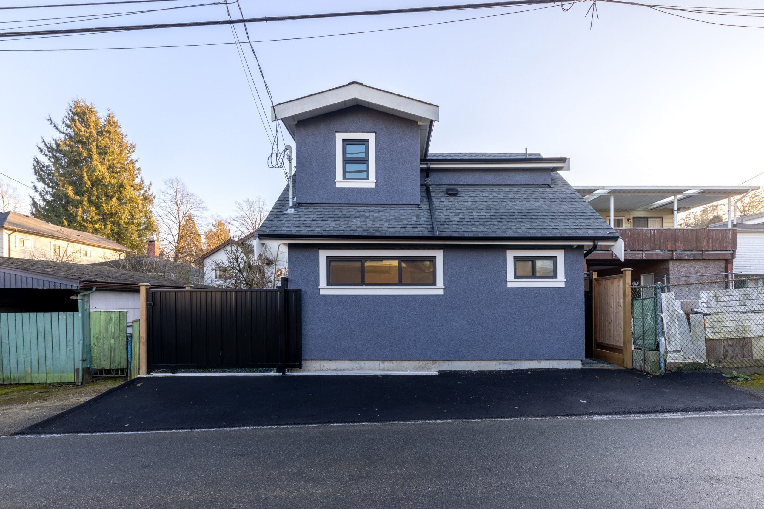 A small modern house with blue exterior walls, a gray roof, and several rectangular windows, seen from the street with a black gate and paved driveway.