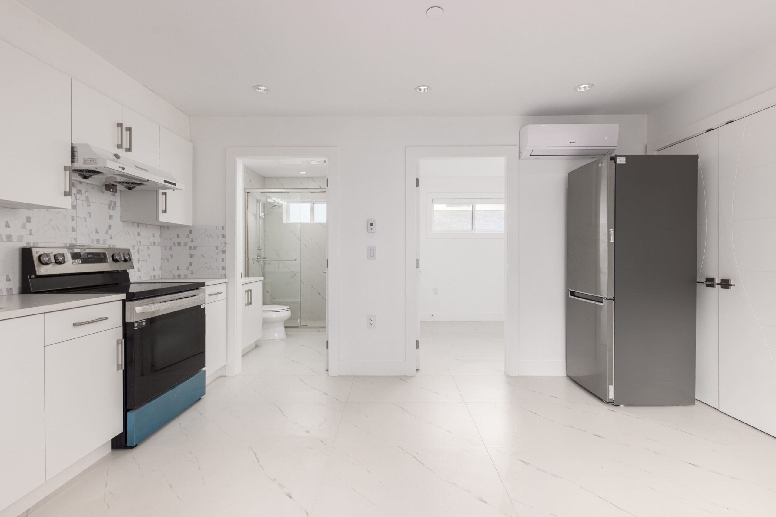 Modern, empty white kitchen with stainless steel appliances, an adjacent bathroom, and a bright adjoining room visible through open doors.