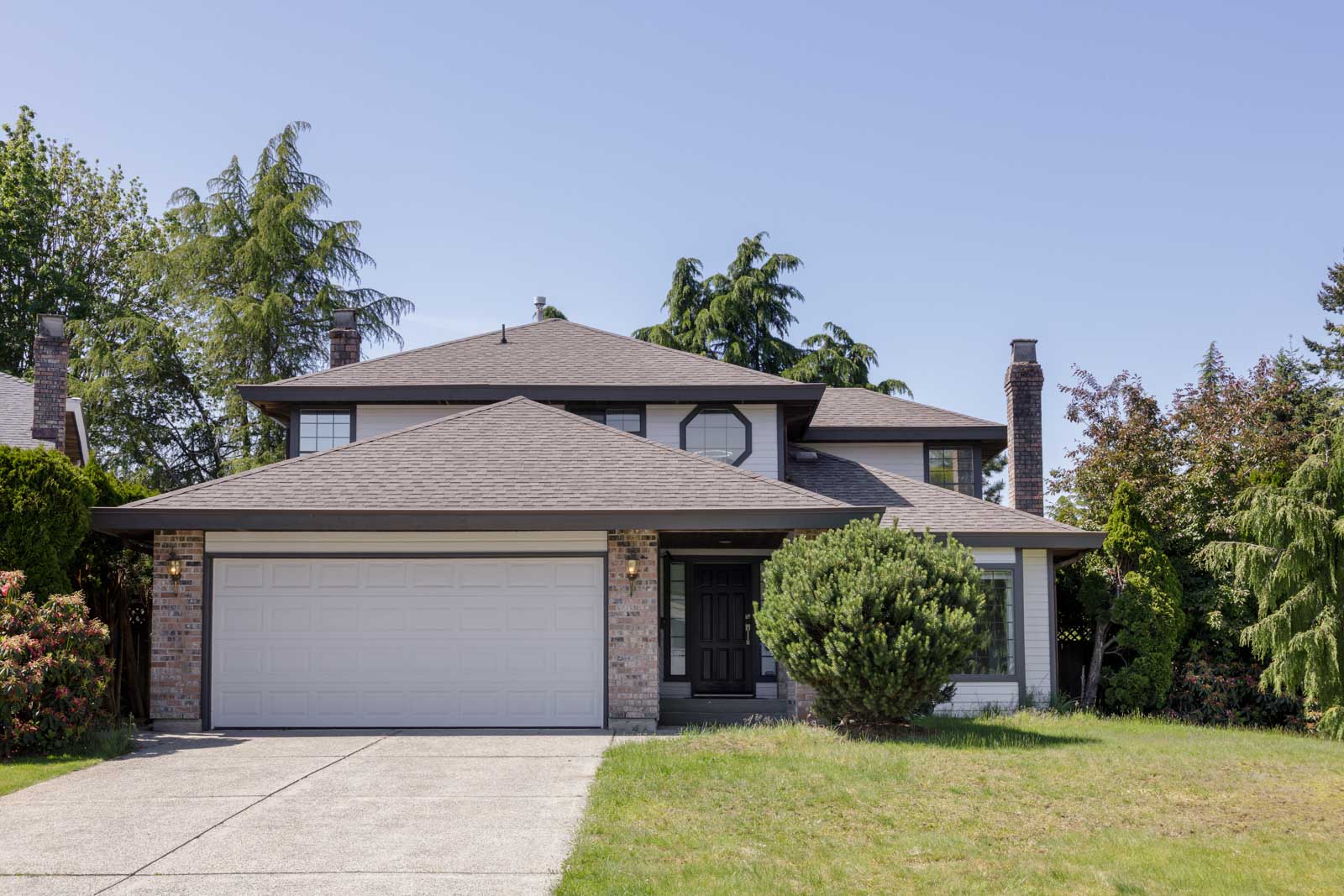 Two-story suburban house with a gray roof, brick and light siding, double garage, black front door, and a driveway, surrounded by trees and a green lawn.