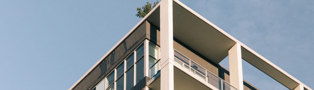 Modern apartment building with glass balconies and a tree visible on the roof against a clear blue sky.