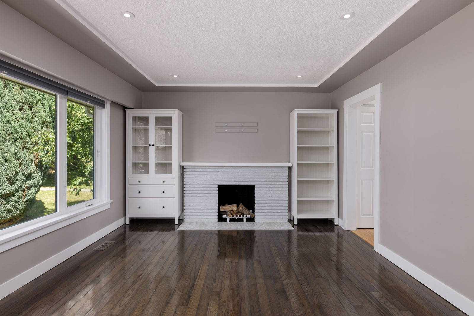 Empty living room with dark wood floors, a white brick fireplace, two white shelving units, and a large window showing greenery outside.