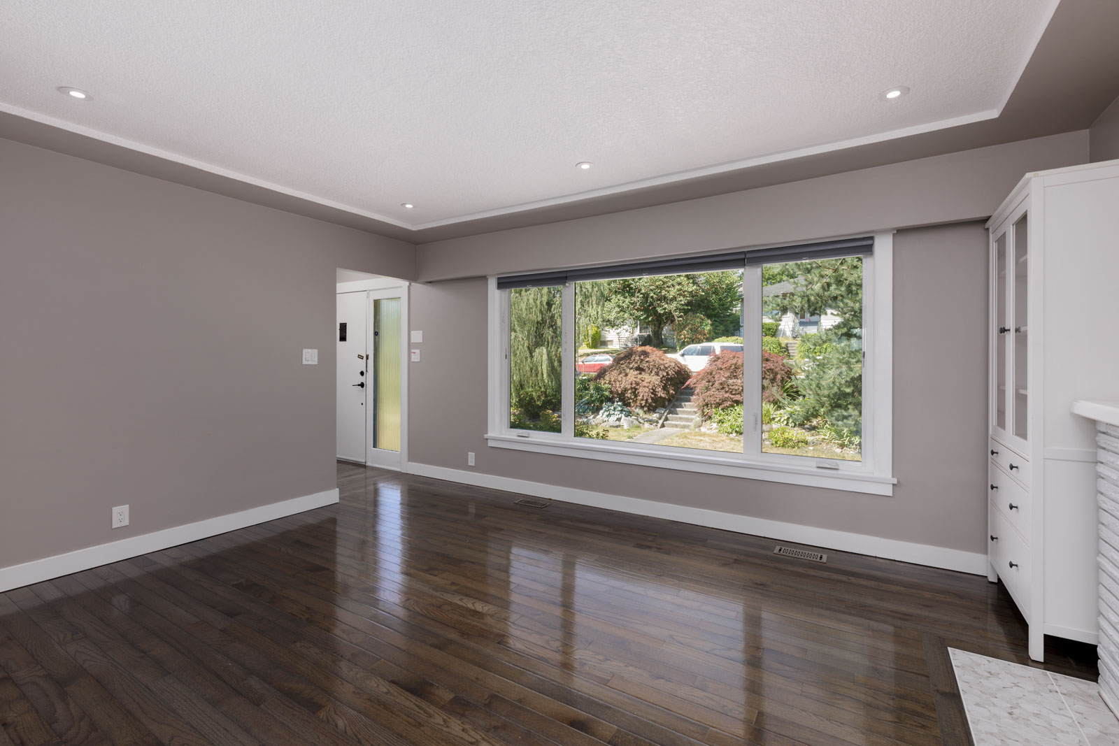 Empty living room with dark hardwood floors, taupe walls, a large front window showing greenery outside, and recessed ceiling lights.