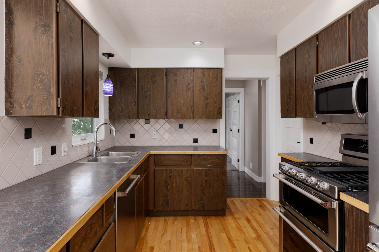 A kitchen with dark wood cabinets, stainless steel appliances, a double sink, and light wood flooring. White tiled backsplash and a small window above the sink.