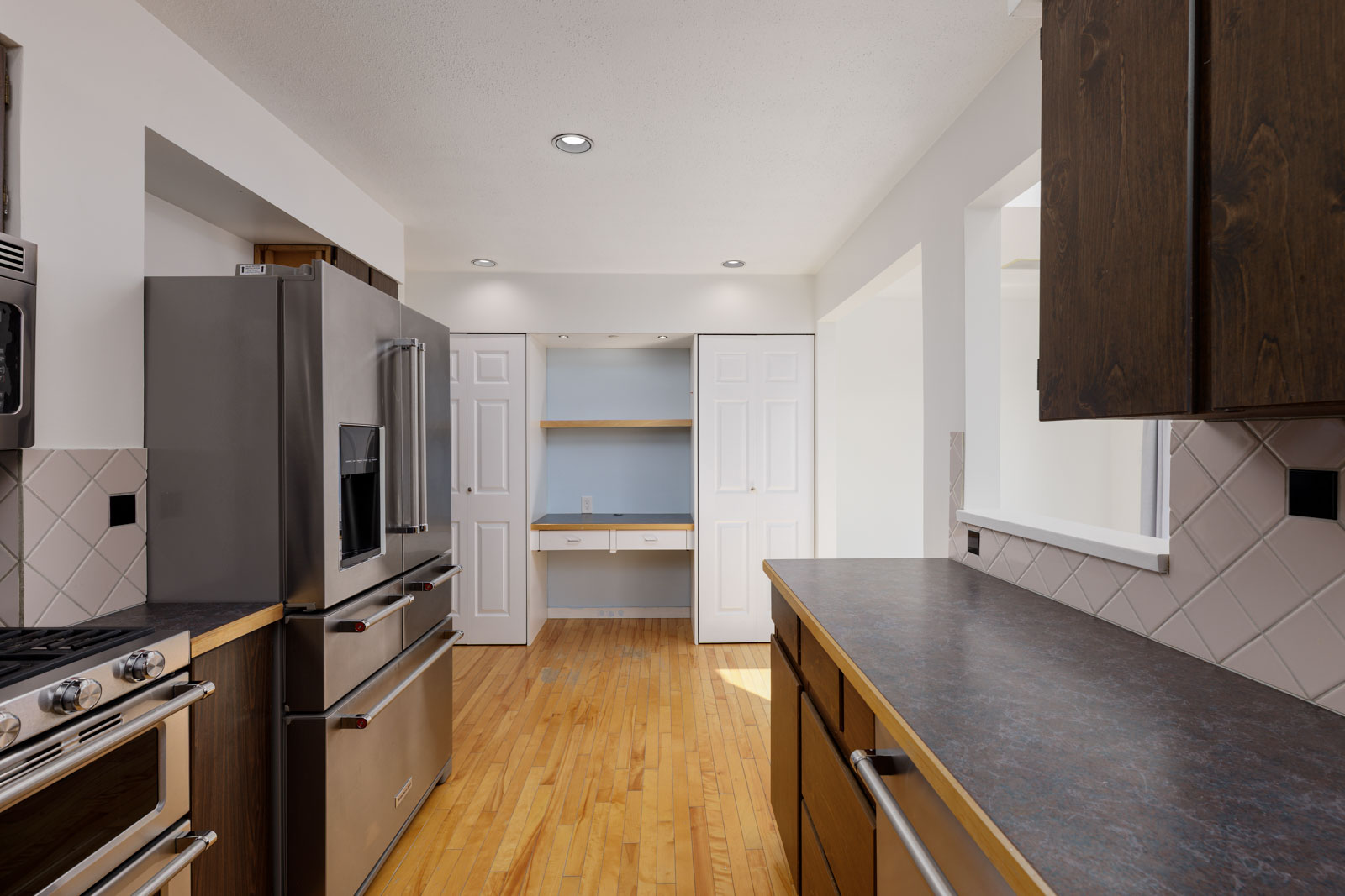 Modern kitchen with stainless steel appliances, wood cabinets, tile backsplash, and a built-in desk area with shelves in an alcove at the far end.