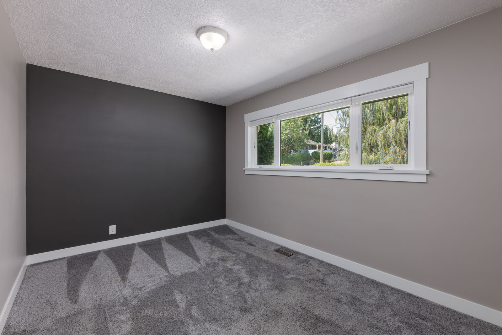 Empty room with gray carpet, a dark accent wall, and a large window letting in natural light. Ceiling light fixture and white baseboards are visible.