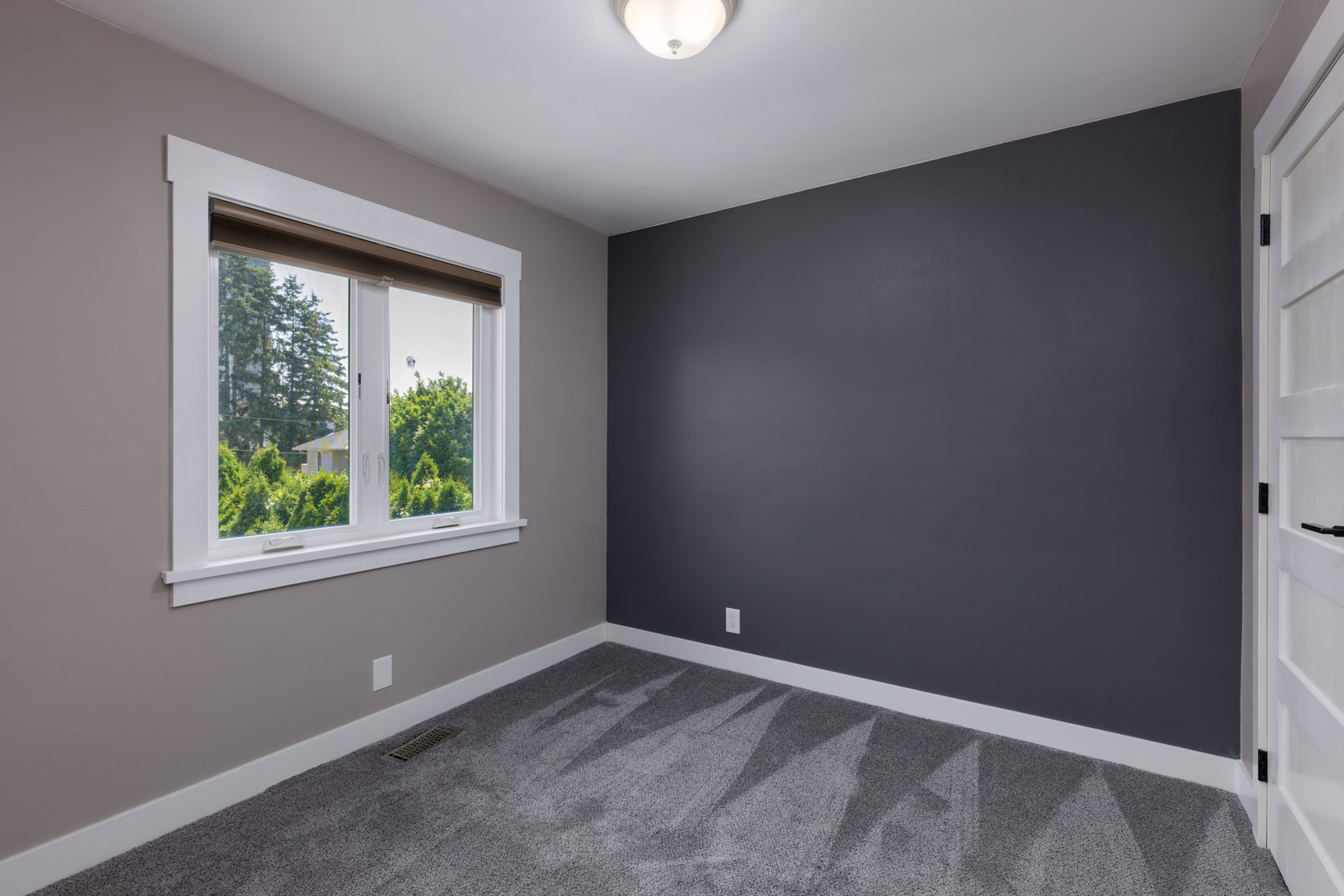 Empty room with gray carpet, a dark accent wall, beige side walls, a white-trimmed window, and a white door, lit by a ceiling light.