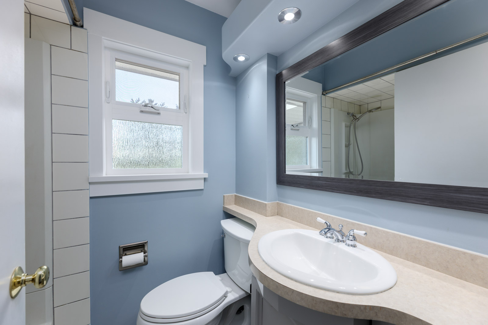 A small bathroom with blue walls, a white sink, a toilet, a large mirror, and a window. The room also features a wall-mounted light above the mirror.