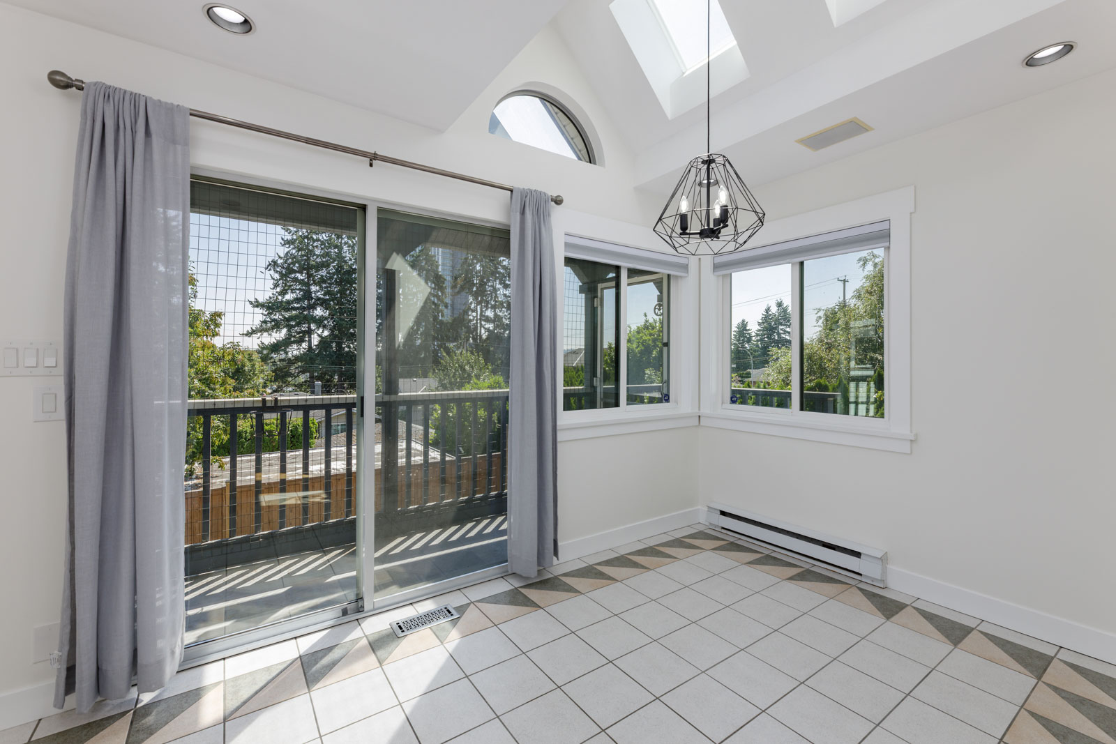 Bright dining area with large windows, skylights, and sliding glass door leading to a balcony; tiled floor and light gray curtains.