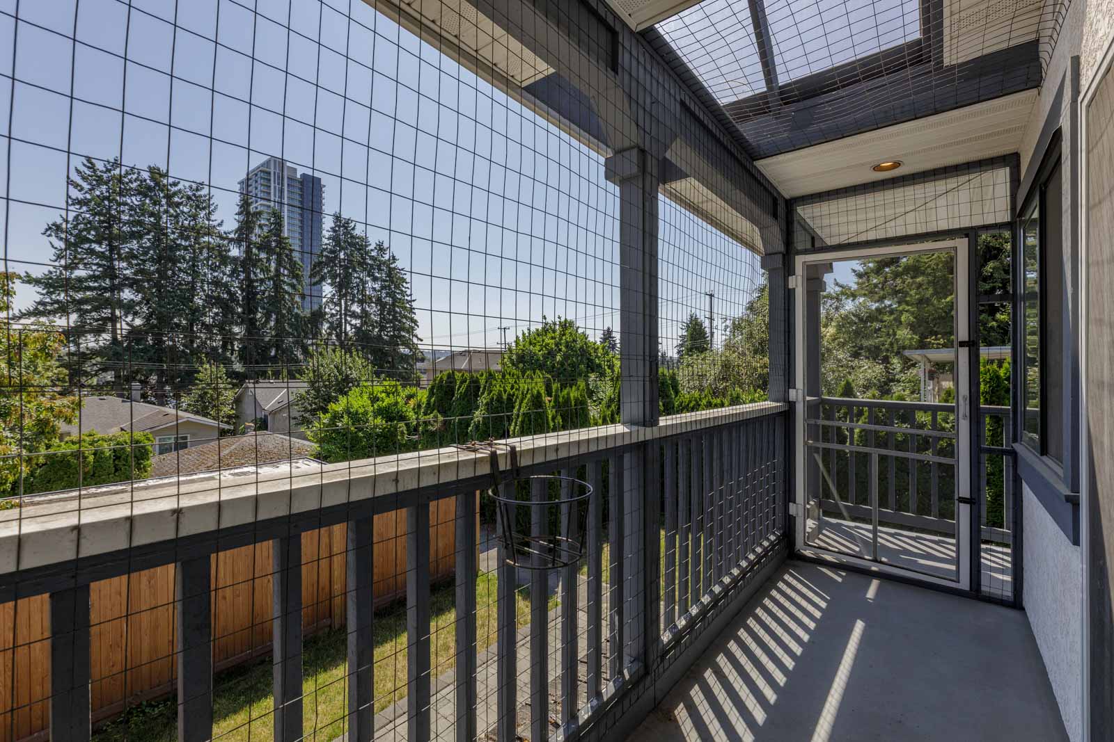 Covered balcony with wire mesh, wooden railing, and a view of trees, houses, and a tall building in the background on a sunny day.
