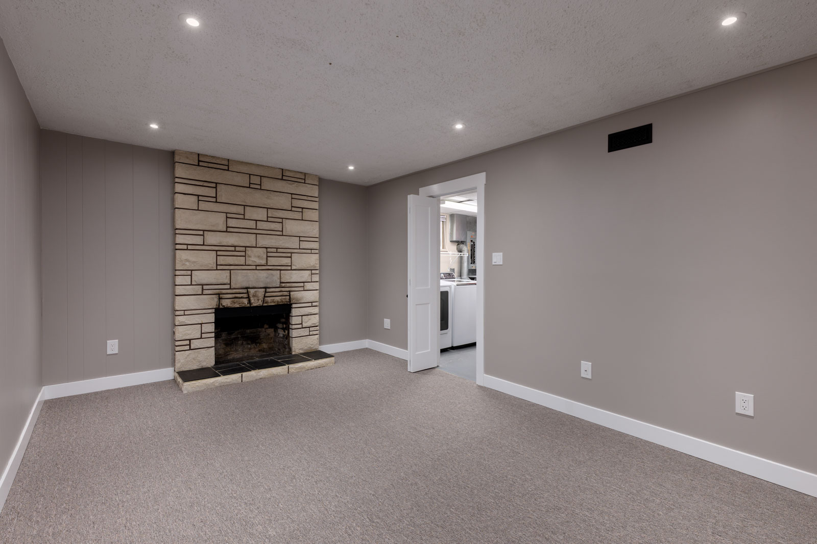 A carpeted room with beige walls, a stone fireplace, recessed ceiling lights, and an open door leading to a kitchen area.
