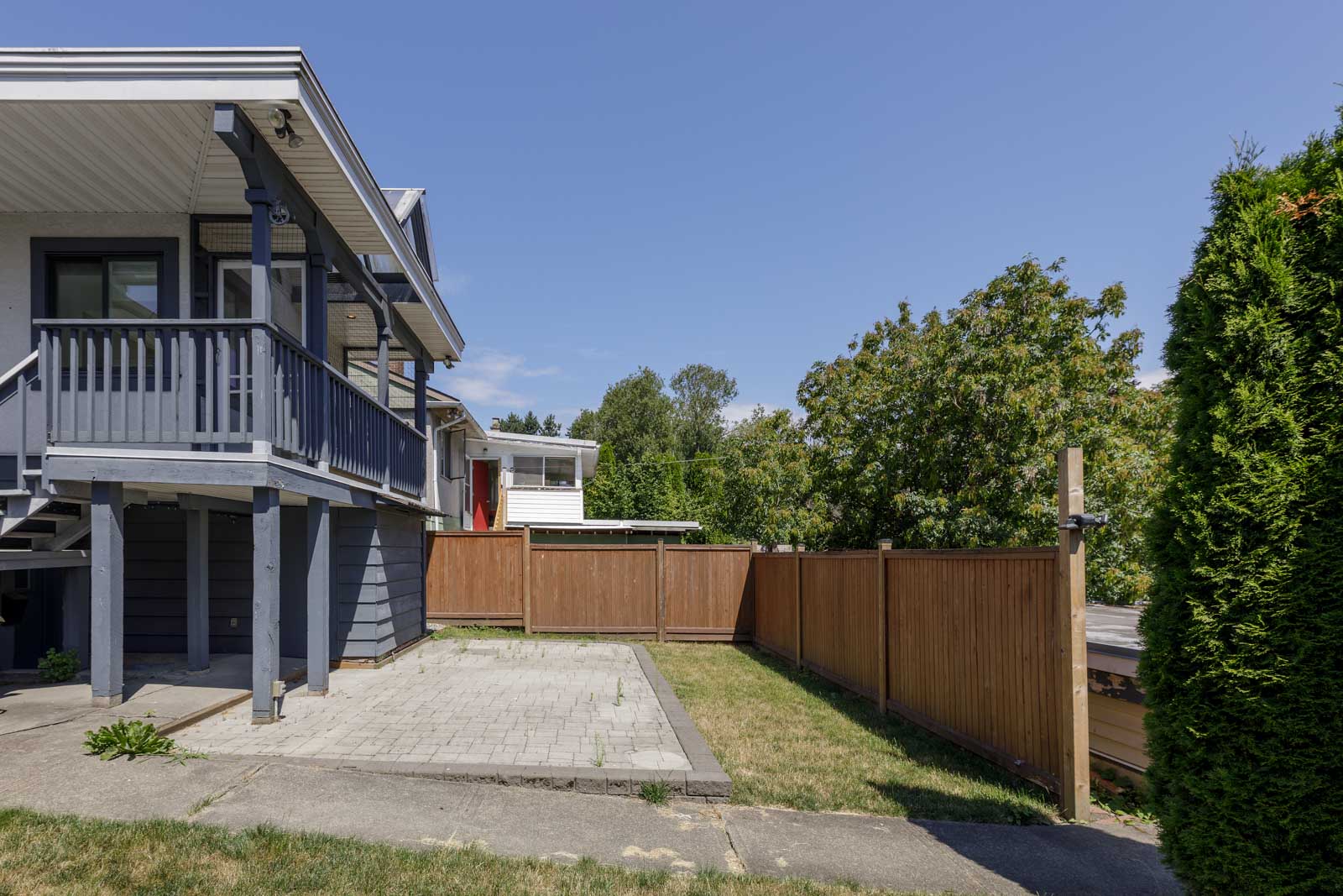 Side view of a house with a raised porch, wooden fence, paved patio area, patchy grass, and trees in the background under a clear sky.