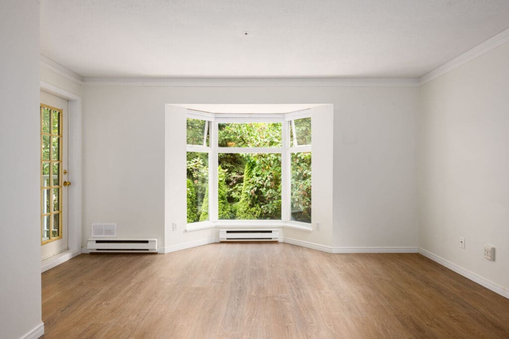 Empty living room with white walls, wood flooring, a large bay window overlooking greenery, and a glass door on the left side.
