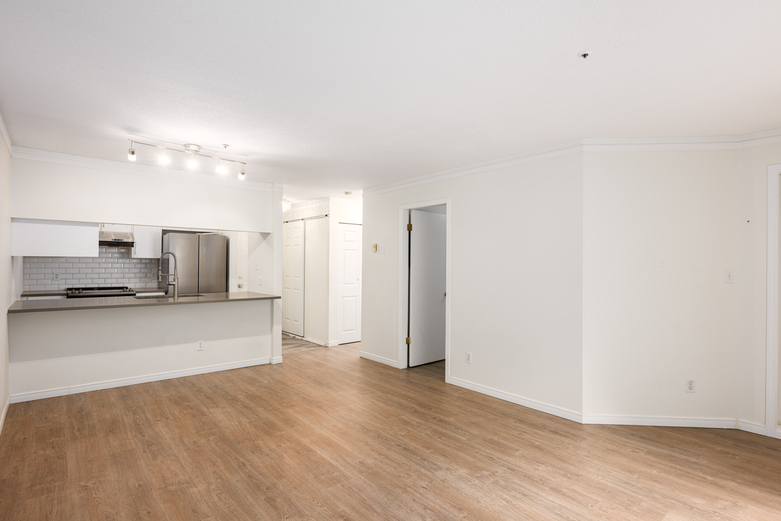 Unfurnished apartment with white walls, wood-look flooring, open kitchen with stainless steel appliances, and visible doorways to other rooms.