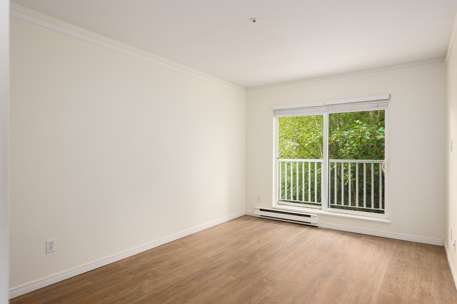 Empty room with light-colored walls, wood flooring, and a large window with white blinds overlooking green trees.