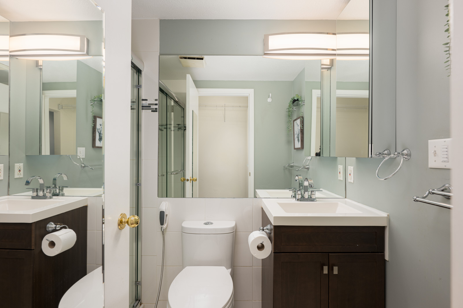 Bathroom with a white toilet, two sinks with dark cabinets, mirrors above each sink, glass shower door, and neutral-colored walls.