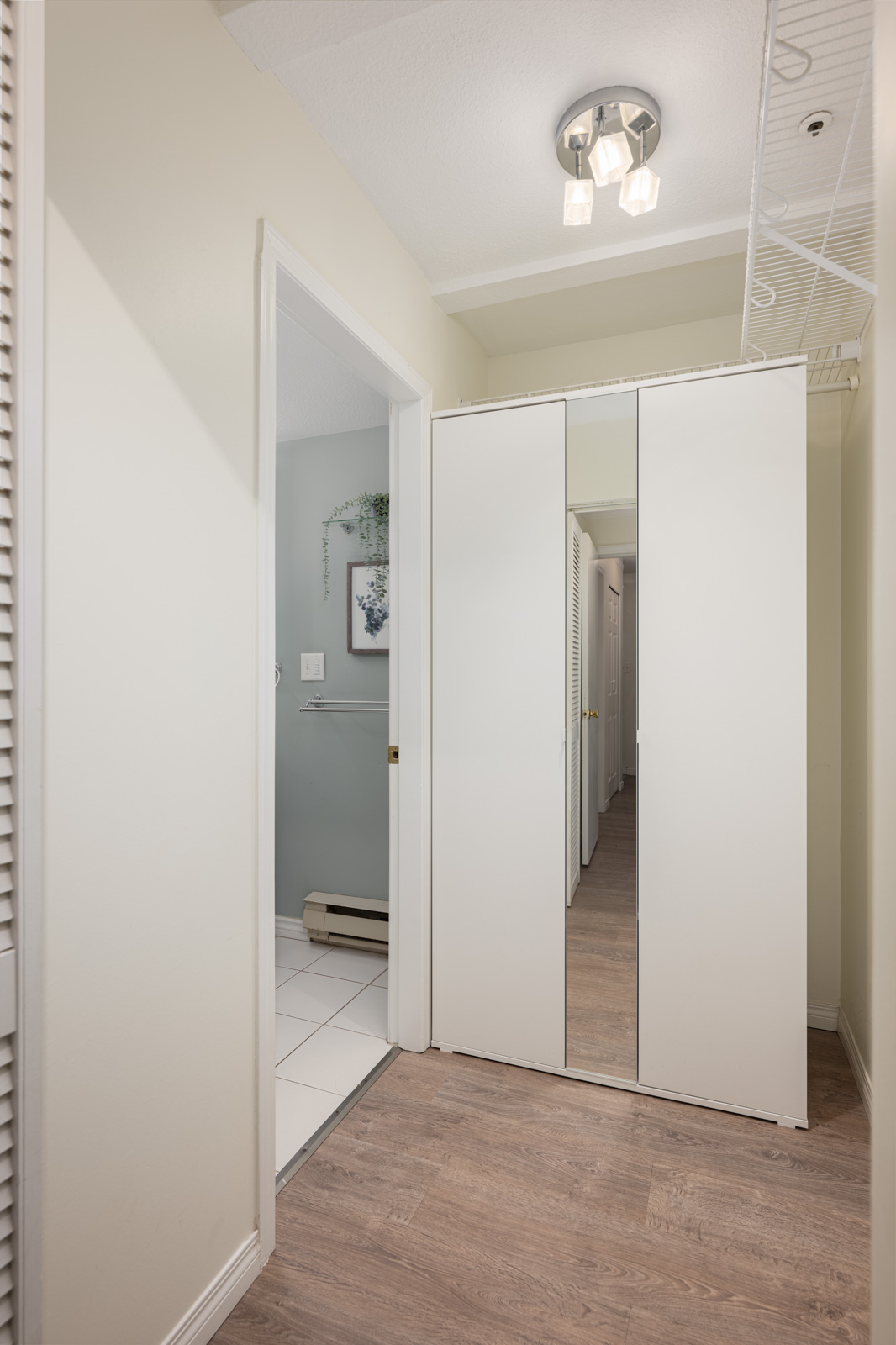 A hallway with light wood flooring, white walls, a ceiling light fixture, a mirrored closet, and a view into a tiled bathroom with framed art on the wall.