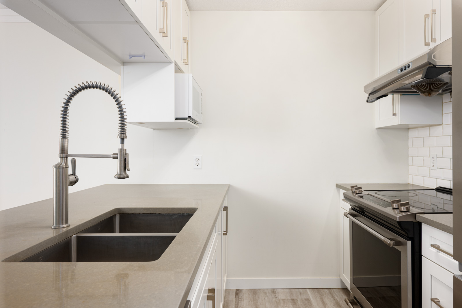 Modern kitchen with a gray countertop, stainless steel sink and faucet, electric stove with oven, white cabinets, and light wood flooring.