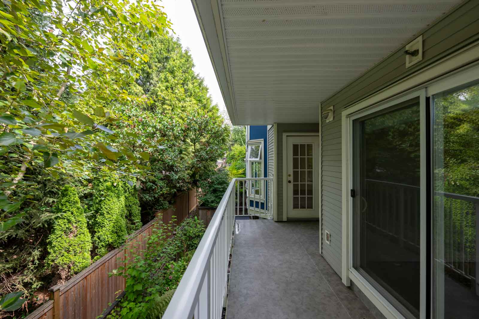 A covered balcony with a white railing overlooks a fenced yard with dense green trees and bushes; a sliding glass door and an exterior door lead inside.