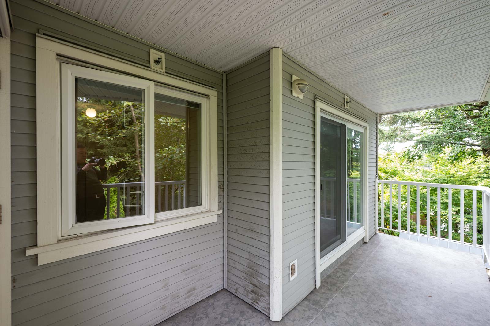 A covered balcony with gray siding, a window, a sliding glass door, white railing, and a view of trees in the background.