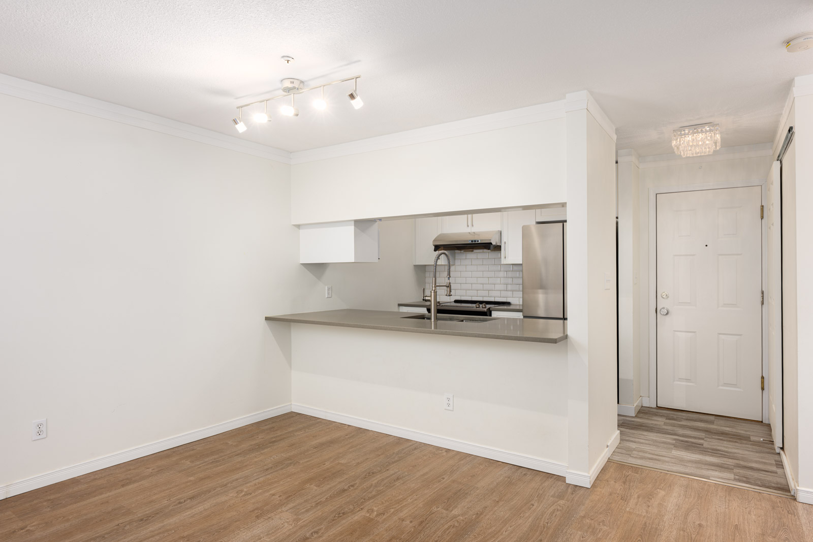 A modern apartment interior with an open kitchen, light wood flooring, white walls, overhead track lighting, and a closed front door in the background.