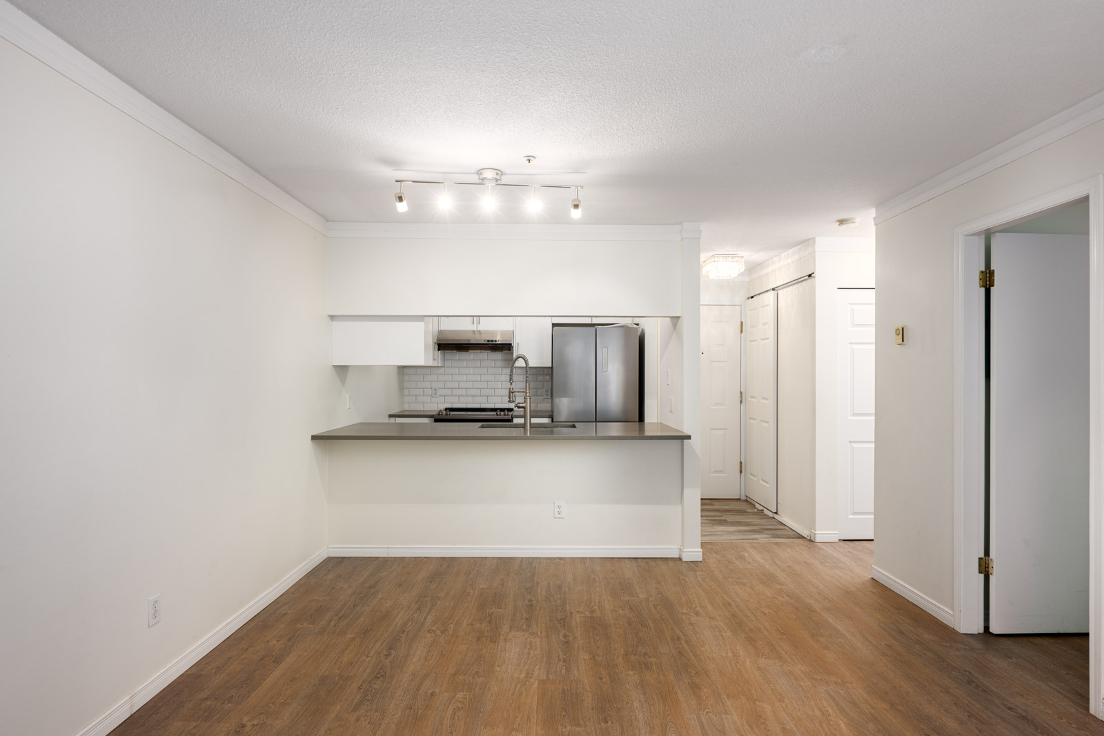 Unfurnished apartment interior with open kitchen, stainless steel appliances, white cabinetry, wood flooring, and white walls.