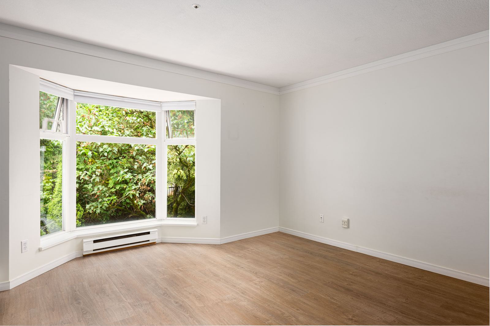 Empty room with light-colored walls, wood flooring, and a large bay window overlooking green foliage.