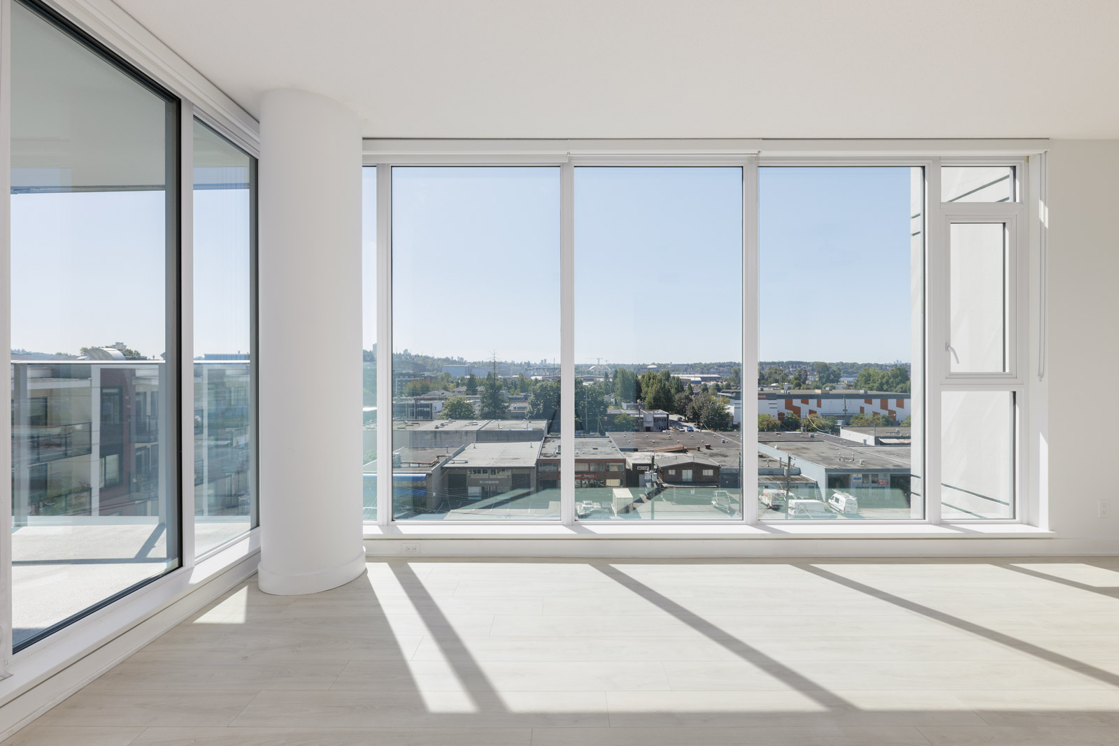 A bright, empty modern room with large floor-to-ceiling windows overlooking a cityscape and blue sky.