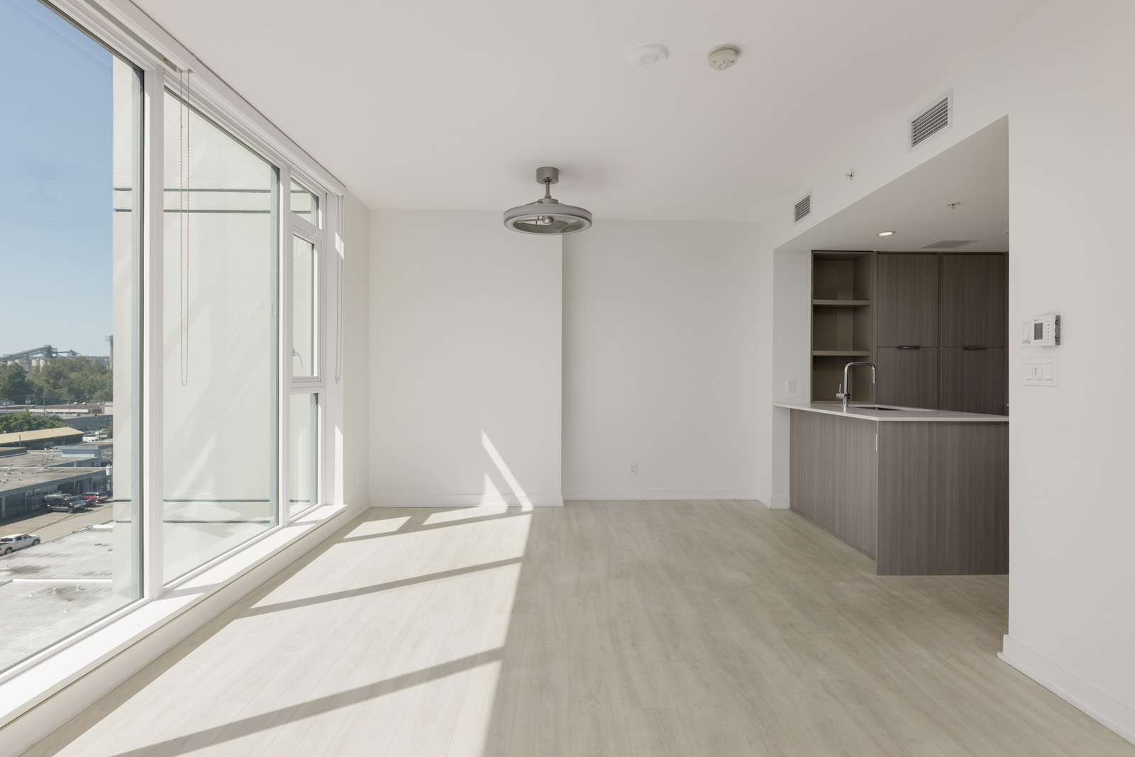 Unfurnished modern apartment with large windows, light wood flooring, white walls, and an open kitchen area with wood cabinets. Sunlight fills the space.
