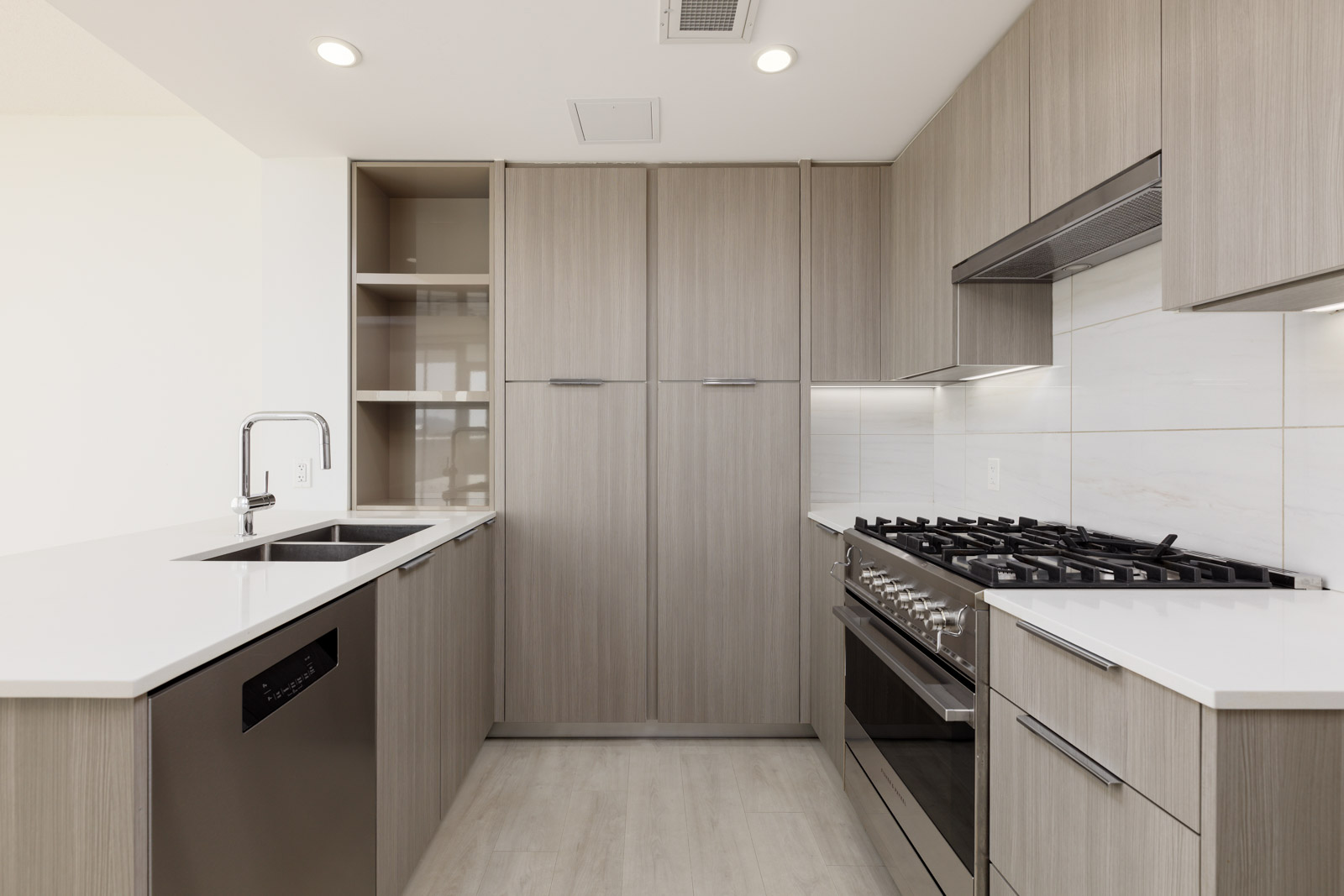 Modern kitchen with light wood cabinets, white countertops, stainless steel appliances, built-in oven, gas stove, and double sink under bright, even lighting.