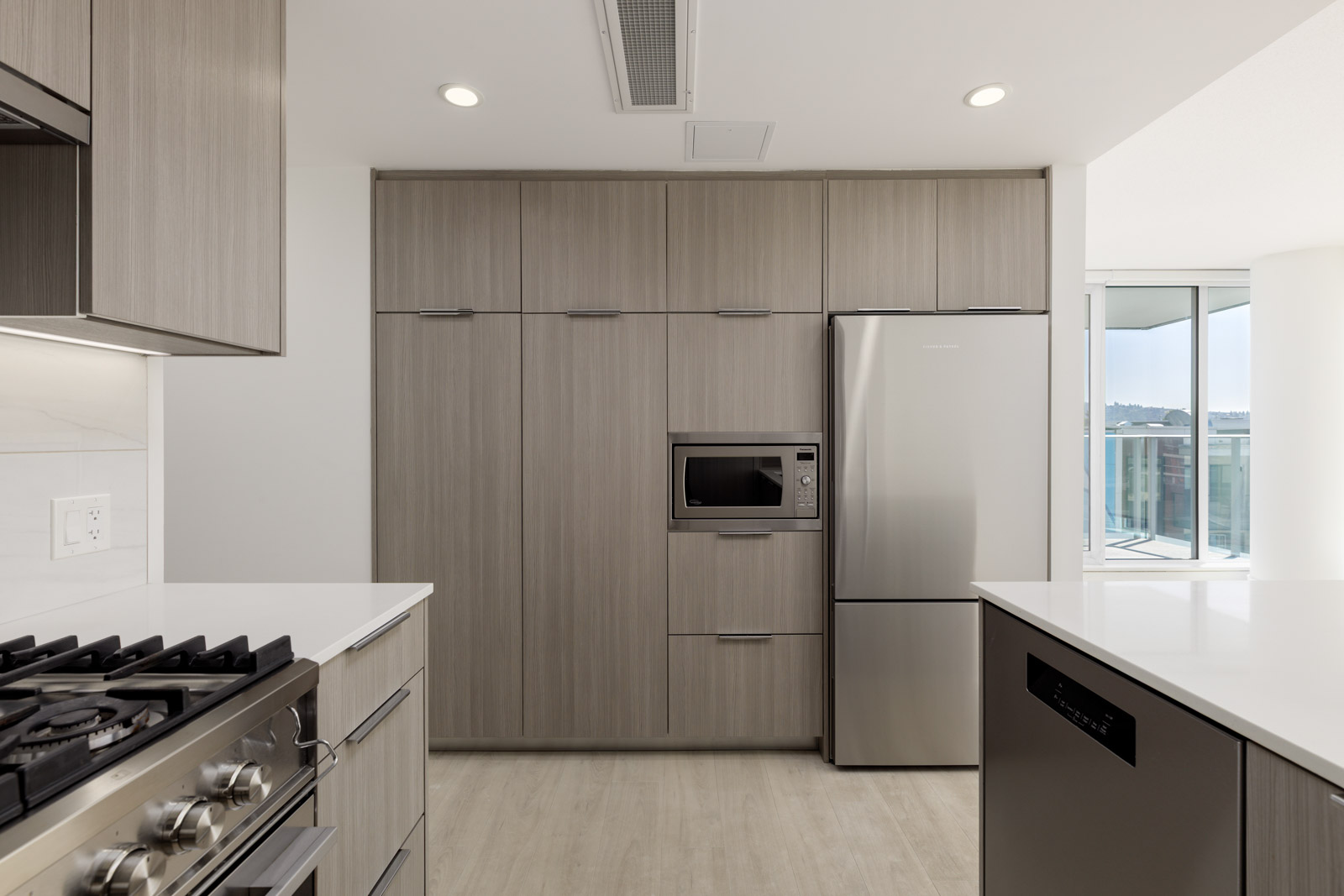 Modern kitchen with light wood cabinets, stainless steel appliances including a refrigerator, microwave, gas stove, and dishwasher, with natural light from large windows.