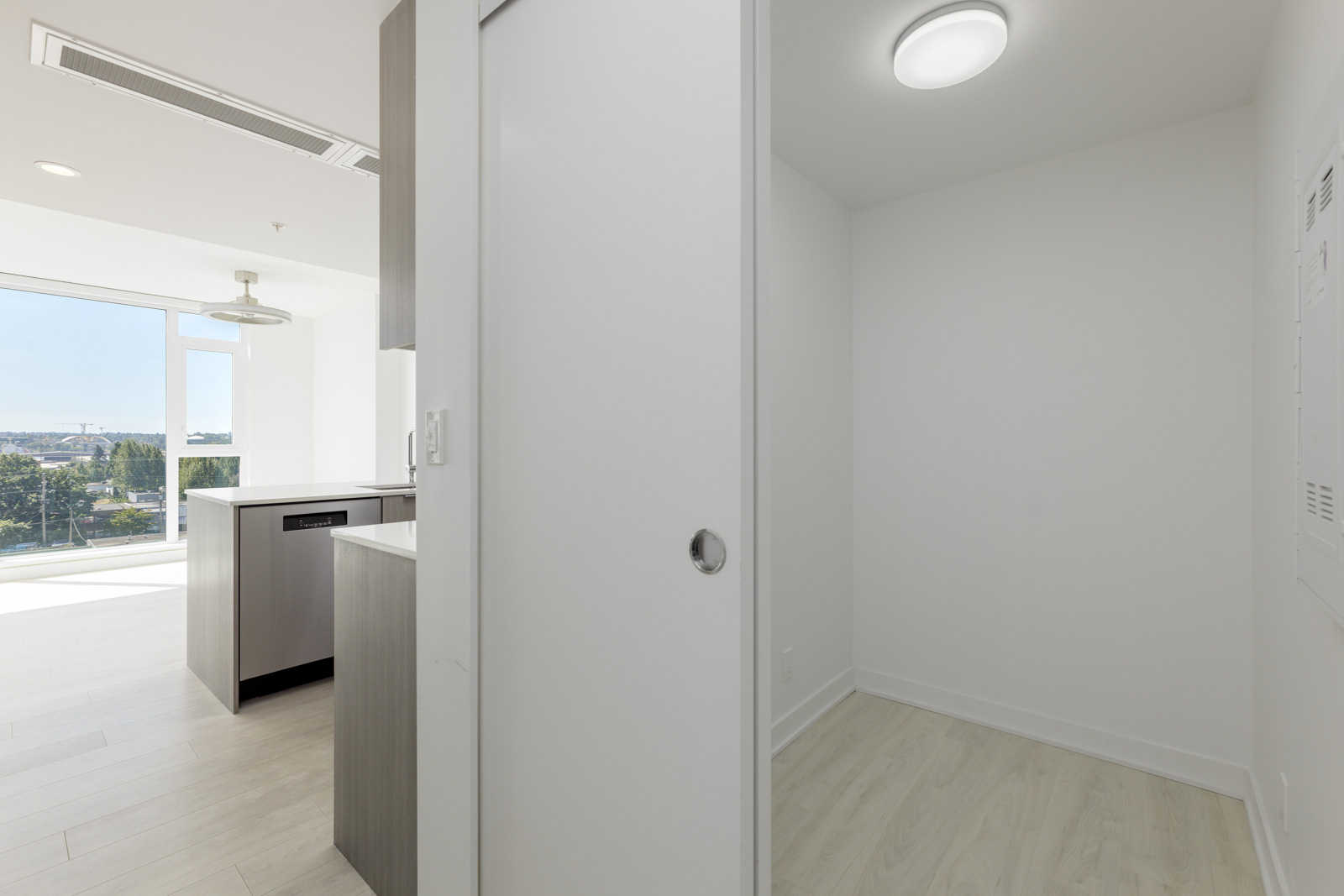 A modern apartment interior with a small empty room, light wood floors, sliding door, and an open kitchen area with large windows in the background.
